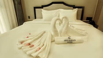 A neatly made bed with a heart-shaped towel arrangement and a rolled towel with 'ANVI SAIGON HOTEL' text.