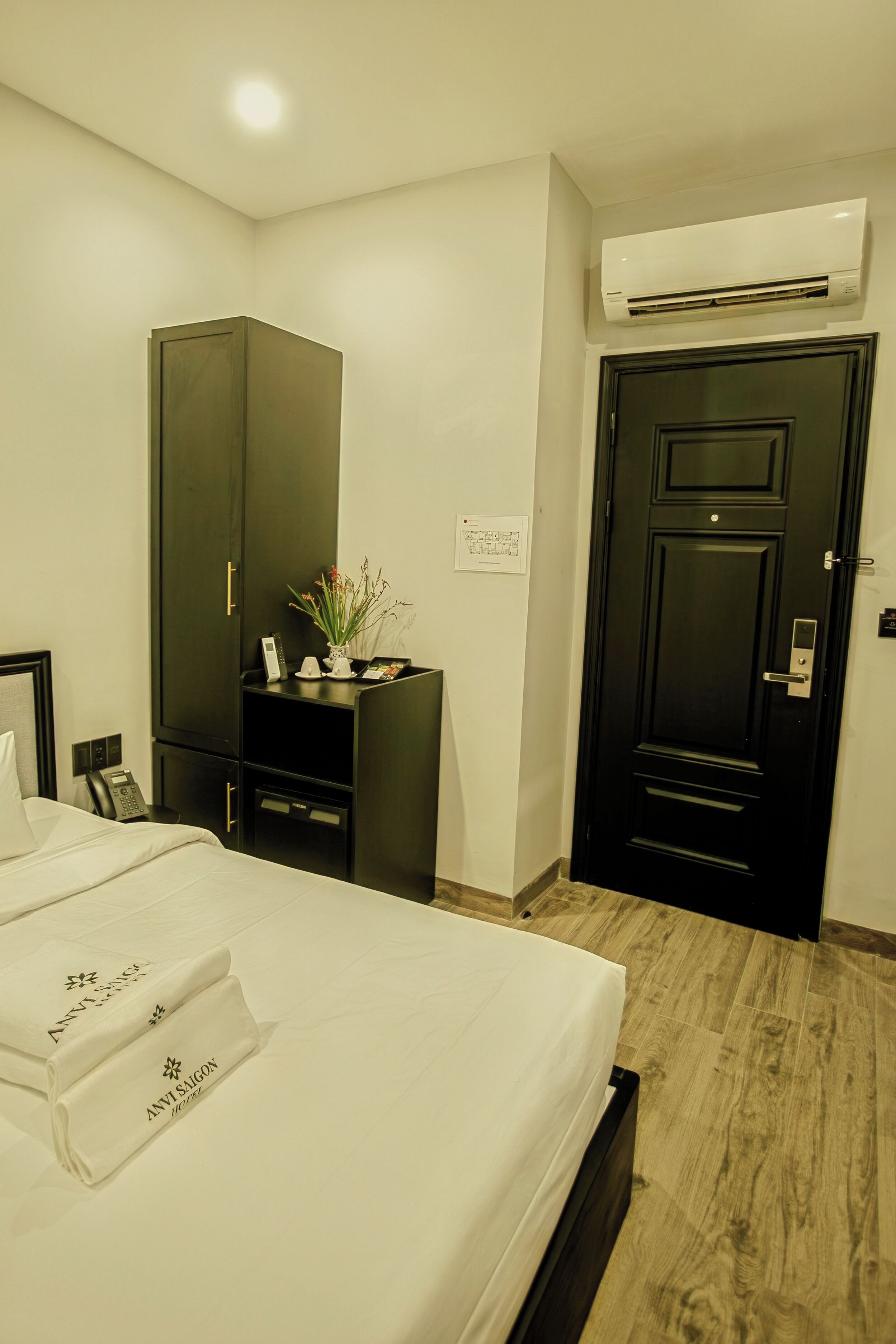 A hotel room with a bed, a wardrobe, a desk with a plant, and an air conditioning unit.