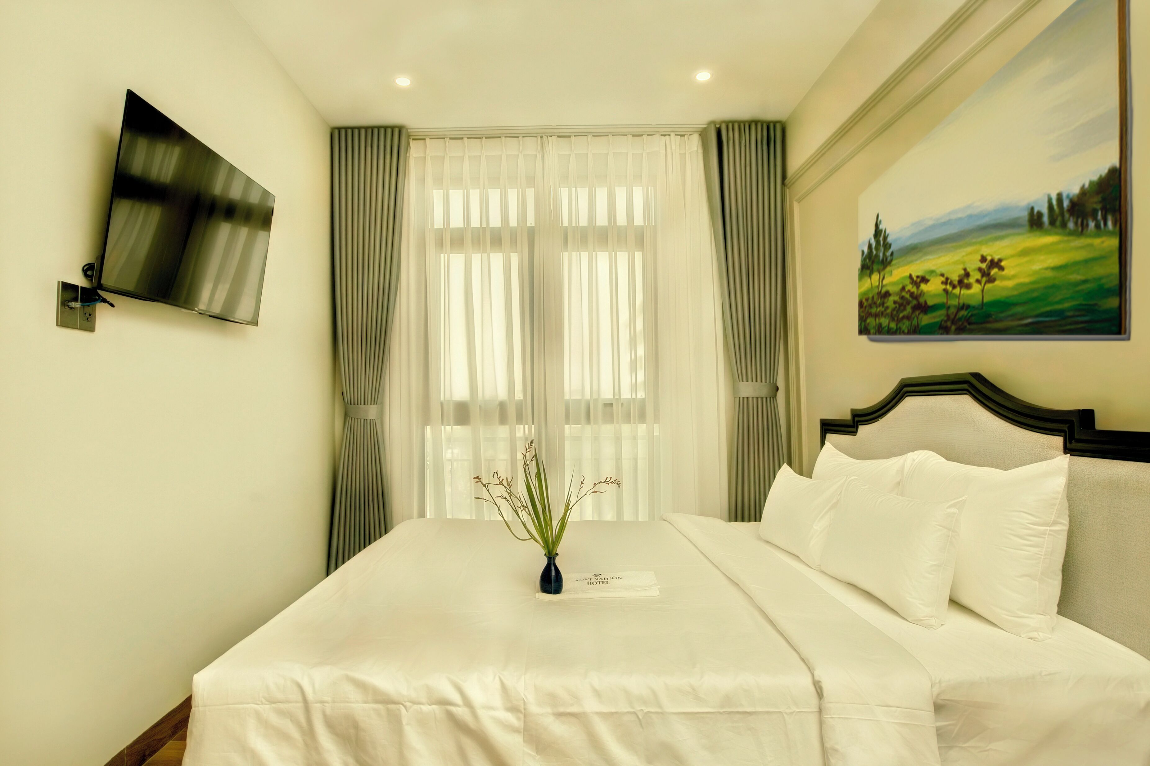 Un lit bien fait, avec du linge de lit blanc, un vase orné de verdure, un tableau accroché au mur et un téléviseur à écran plat fixé au mur.