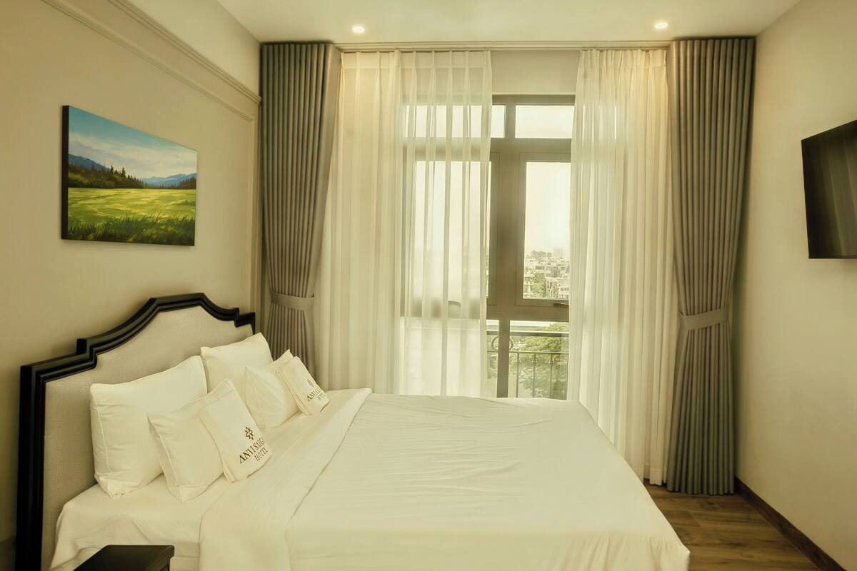 A hotel room with a large bed, a painting on the wall, and a view through the window.