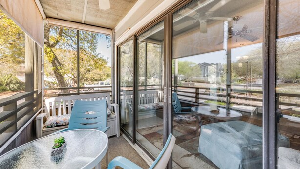 Un patio techado al aire libre con pared de vidrio, muebles blancos y una mesa pequeña con una planta.