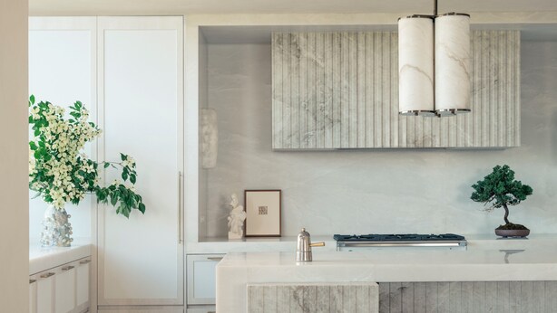 Une cuisine moderne avec un plan de travail en marbre, deux tabourets, un luminaire suspendu et une plante en pot.