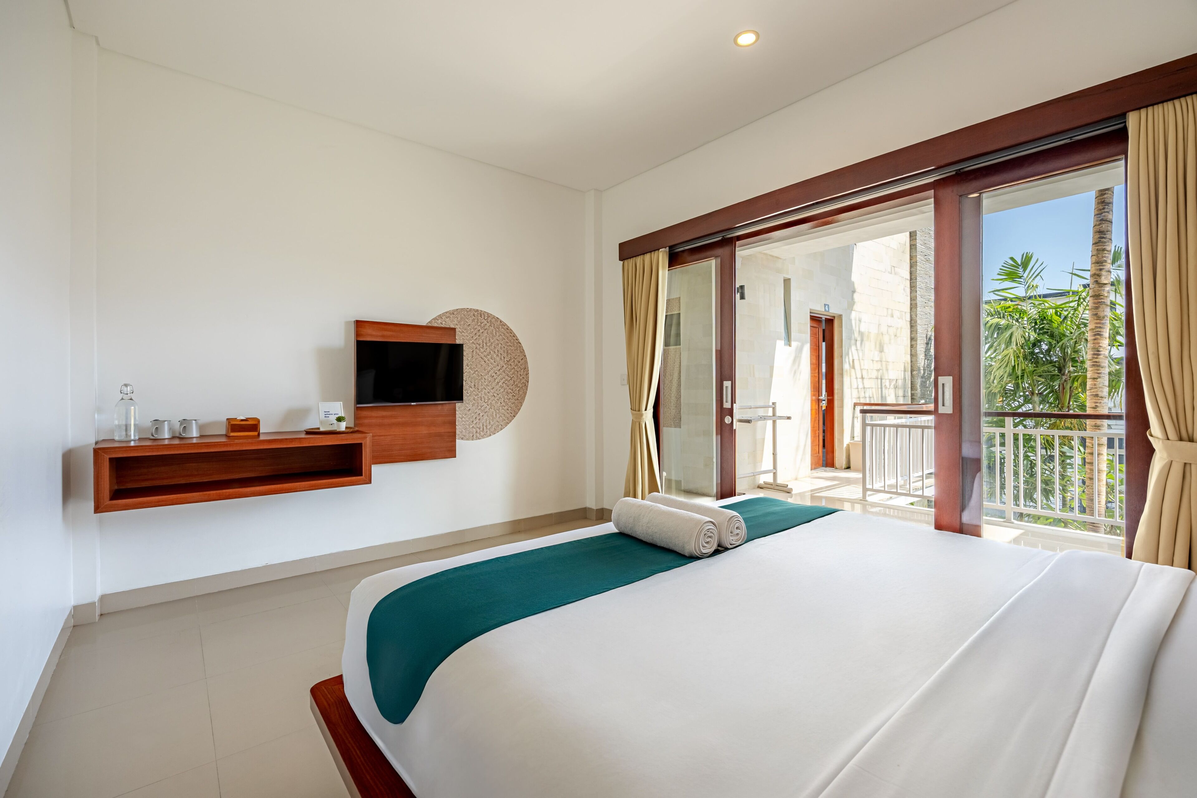 Habitación de hotel con cama, televisor montado, estante de madera con artículos de aseo y balcón con vistas a palmeras.