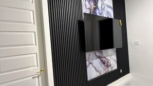 Una habitación moderna con una puerta blanca, un televisor de pantalla plana montado sobre un panel negro con una obra de arte que imita el mármol, y un piso de baldosas blancas.