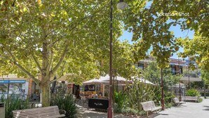 Un área de estar al aire libre con mesas y bancos bajo árboles, una farola y un edificio con señalización al fondo.