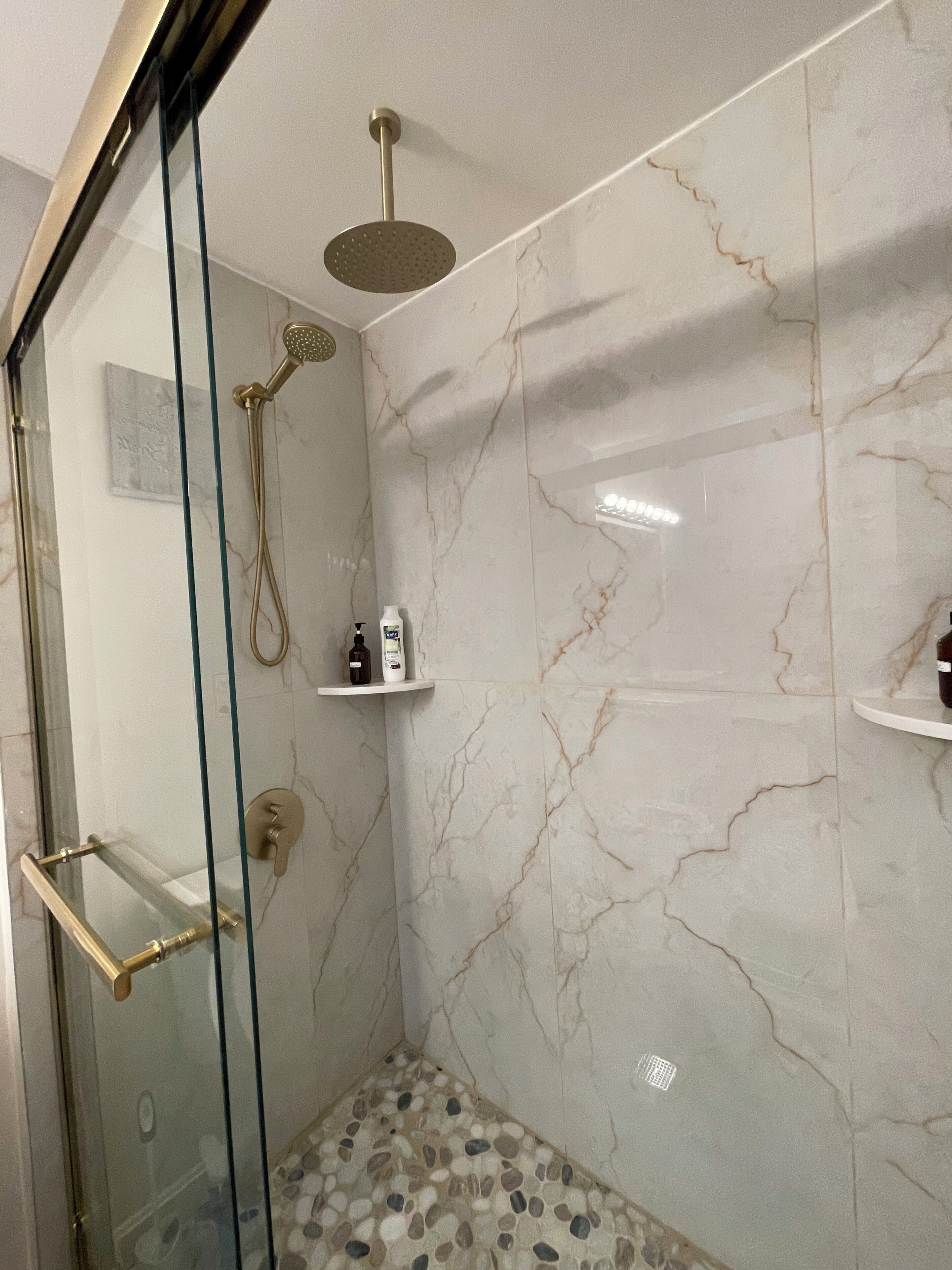 Een glazen douchecabine met een handdouche en een douchekop aan de muur. Er zijn planken met toiletartikelen en een vloer met een marmerlook.