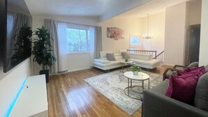 Un salon avec un canapé, une table basse et un téléviseur. Il y a une fenêtre avec des rideaux, une plante et un lustre.