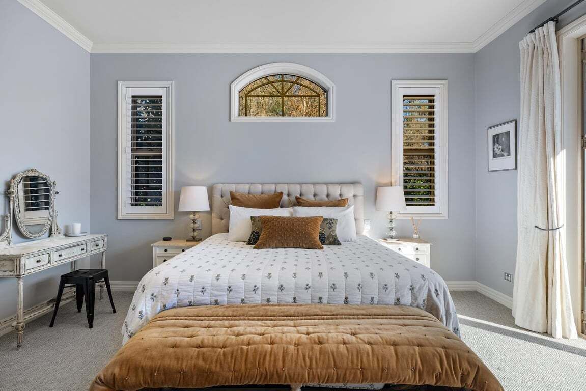 A bedroom with a large bed, two bedside tables, a dresser, a mirror, and two windows with shutters.