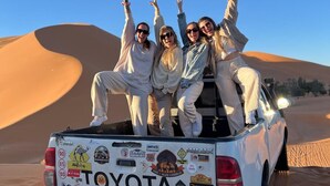 Una camioneta Toyota blanca con cuatro pasajeros, algunos vestidos de blanco, estacionada sobre una duna de arena con un cielo azul despejado al fondo.