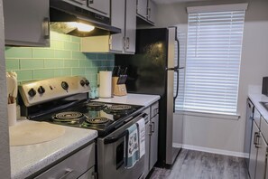 Una cocina con estufa, horno y refrigerador. Hay armarios, una ventana con persianas y una encimera con un bloque de cuchillos y un rollo de toallas de papel.