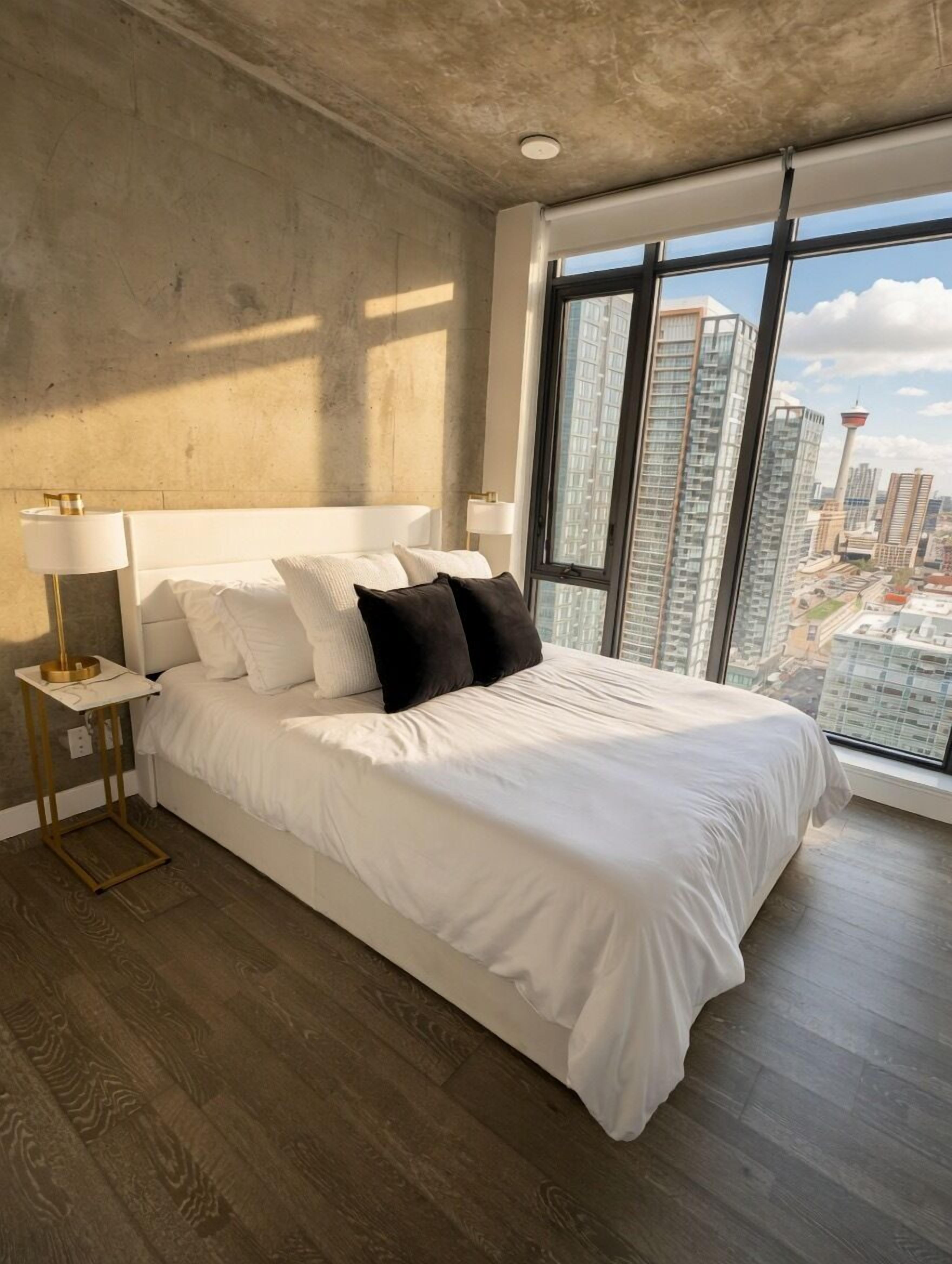 A modern bedroom with a large bed, a nightstand, a window with a city view, and a concrete ceiling.
