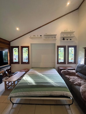 A bedroom with a bed, a TV, a sofa, and two windows with stained glass.