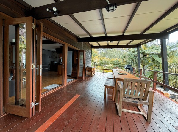 A wooden deck with a dining table and chairs, a grill, and a view of trees and foliage.