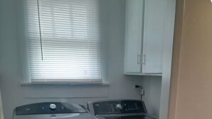 A laundry area with a front-loading washer and dryer, a wooden floor, and a window with blinds.