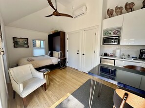 Une pièce de taille modeste comprenant un lit, un fauteuil blanc, une petite table, un coin cuisine et un ventilateur de plafond.