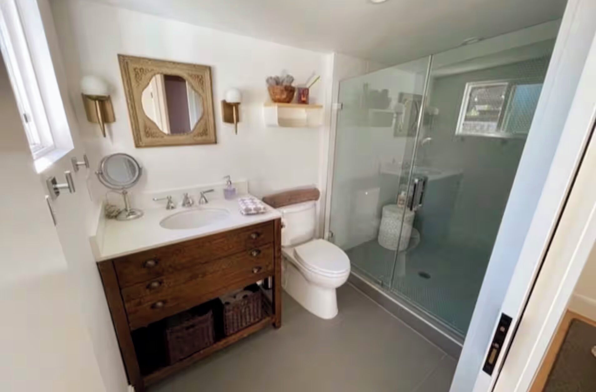 A bathroom with a wooden vanity, a round mirror, a toilet, and a glass shower enclosure.