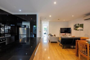 A modern kitchen with a black countertop and stainless steel appliances, a wooden dining area with a table and chairs, and a living space with a sofa and a flat-screen TV. - Puddings Home 4BR Bundoor modern stay family (Bundoora)