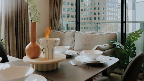 A dining area with a table set for two, a vase with a plant, and a view of a cityscape through large windows. - 5 Min to Crown & NGV | Stylish 2BR | City View (Southbank)