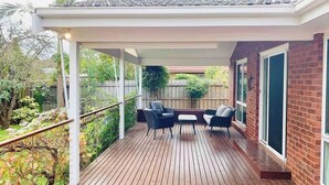 A covered patio with a wooden deck, a table with chairs, and a brick wall with large windows. - Puddings Home Wheelers Hill A/C WiFi Garden (Wheelers Hill)
