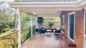 Eine überdachte Terrasse mit Holzdeck, einem Tisch mit Stühlen und einer Ziegelwand mit großen Fenstern.