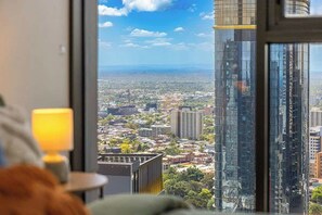 A room with a large window offering a view of a cityscape and a tall building. - Puddings Home Designer City Views & Balcony (Melbourne)