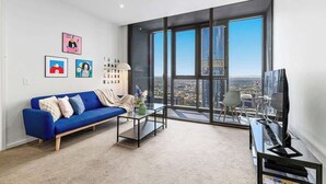 Un moderno salón con un sofá azul, una mesa de centro y un televisor. Amplias ventanas ofrecen vistas a la ciudad.