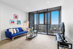 A modern living room with a blue sofa, a coffee table, and a TV. Large windows offer a city view. - Puddings Home Designer City Views & Balcony (Melbourne)