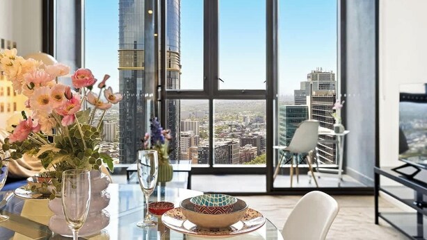 Una habitación de hotel moderna con un ventanal que ofrece vistas a la ciudad, una mesa de comedor con un jarrón de flores y una mesa de vidrio con cristalería.