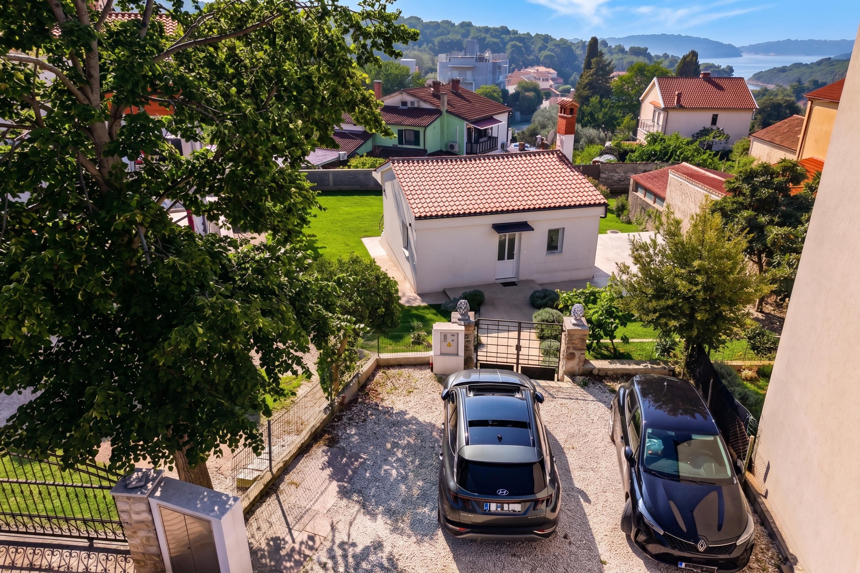 A residential area with two parked cars, a house with a red-tiled roof, and a view of a lake and hills in the background.