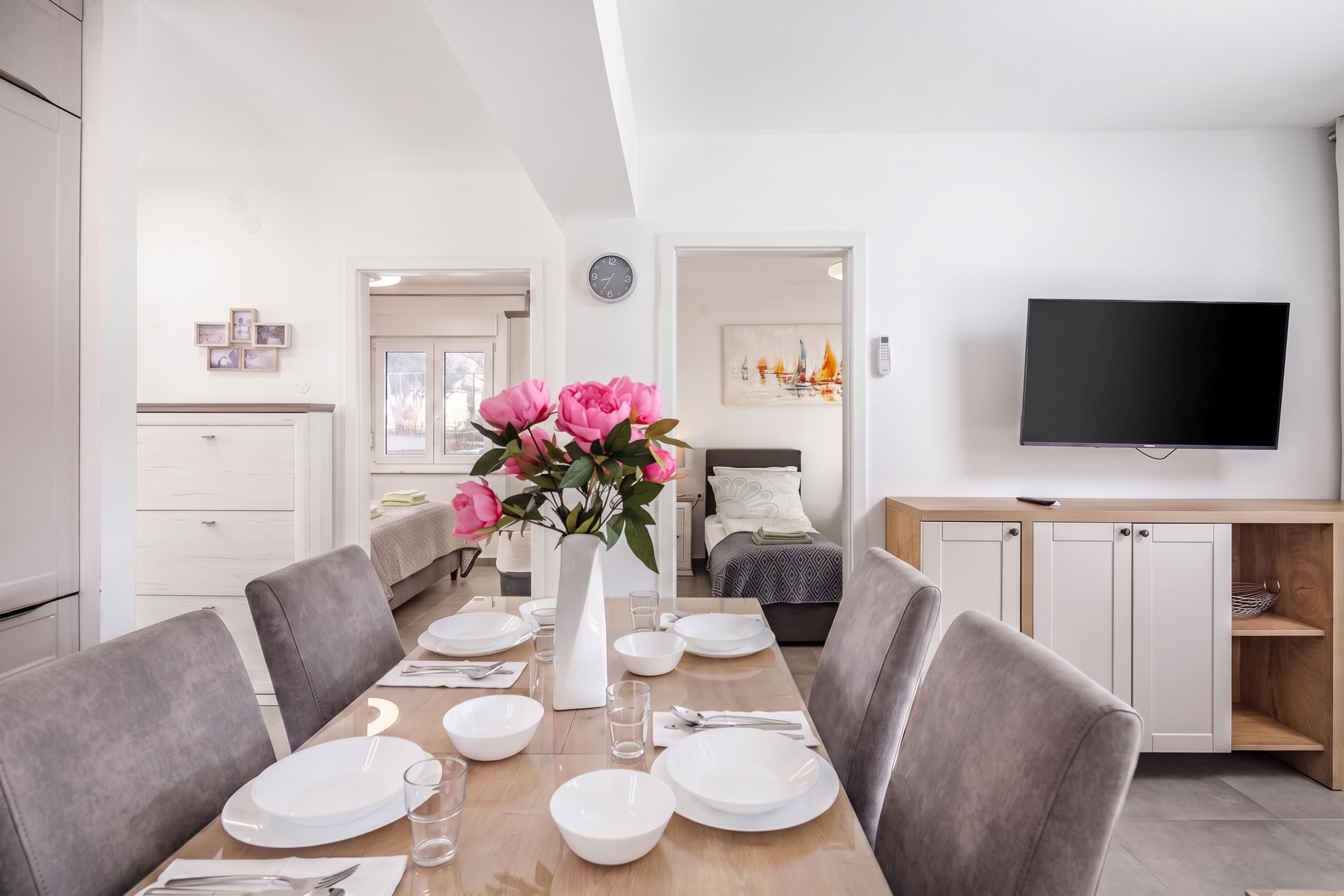 Un espace repas moderne avec une table en bois dressée pour quatre personnes, un vase de fleurs et un téléviseur à écran plat fixé au mur.