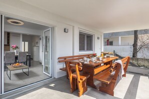Un espace repas moderne avec une table et des bancs en bois, une porte vitrée donnant sur la cuisine et une vue sur un arbre à l’extérieur.