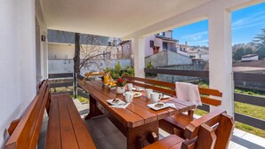 A wooden table set with a bottle, glasses, and a plate with food, surrounded by benches, with a view of buildings and greenery. - Susi's Holiday Home (Medulin)