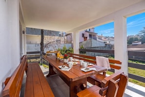 A wooden table set with a bottle, glasses, and a plate with food, surrounded by benches, with a view of buildings and greenery. - Susi's Holiday Home (Medulin)
