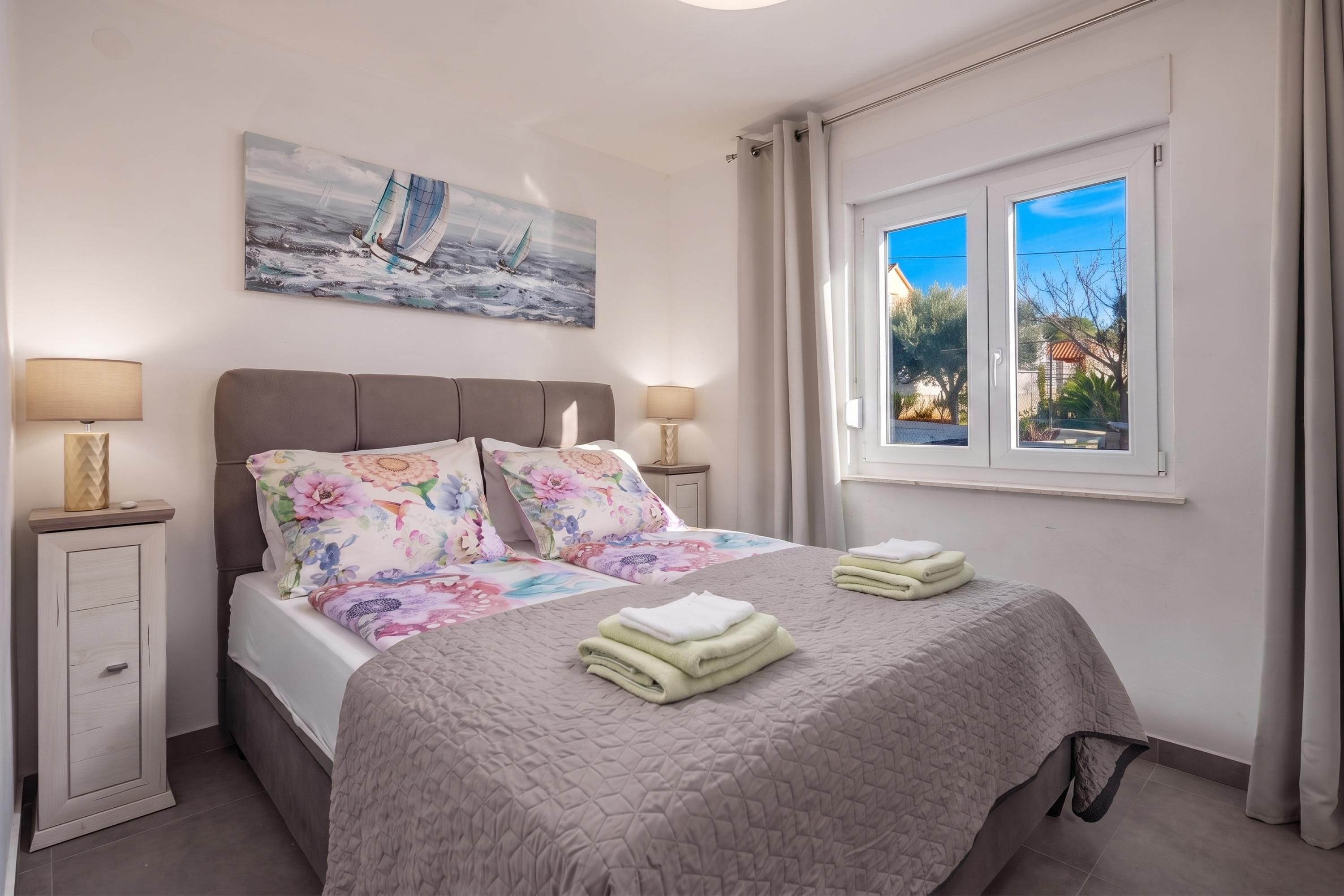 A bedroom with a bed, two bedside tables, a painting, a window with curtains, and a view of the outside.