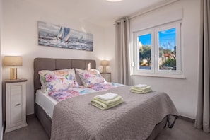 Une chambre à coucher avec un lit, deux tables de chevet, un tableau, une fenêtre avec des rideaux et une vue sur l’extérieur.