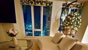 Una sala de estar con una mesa de centro redonda de vidrio, un sofá color beige con cojines, un jarrón con flores y una ventana con vista a la ciudad de noche.
