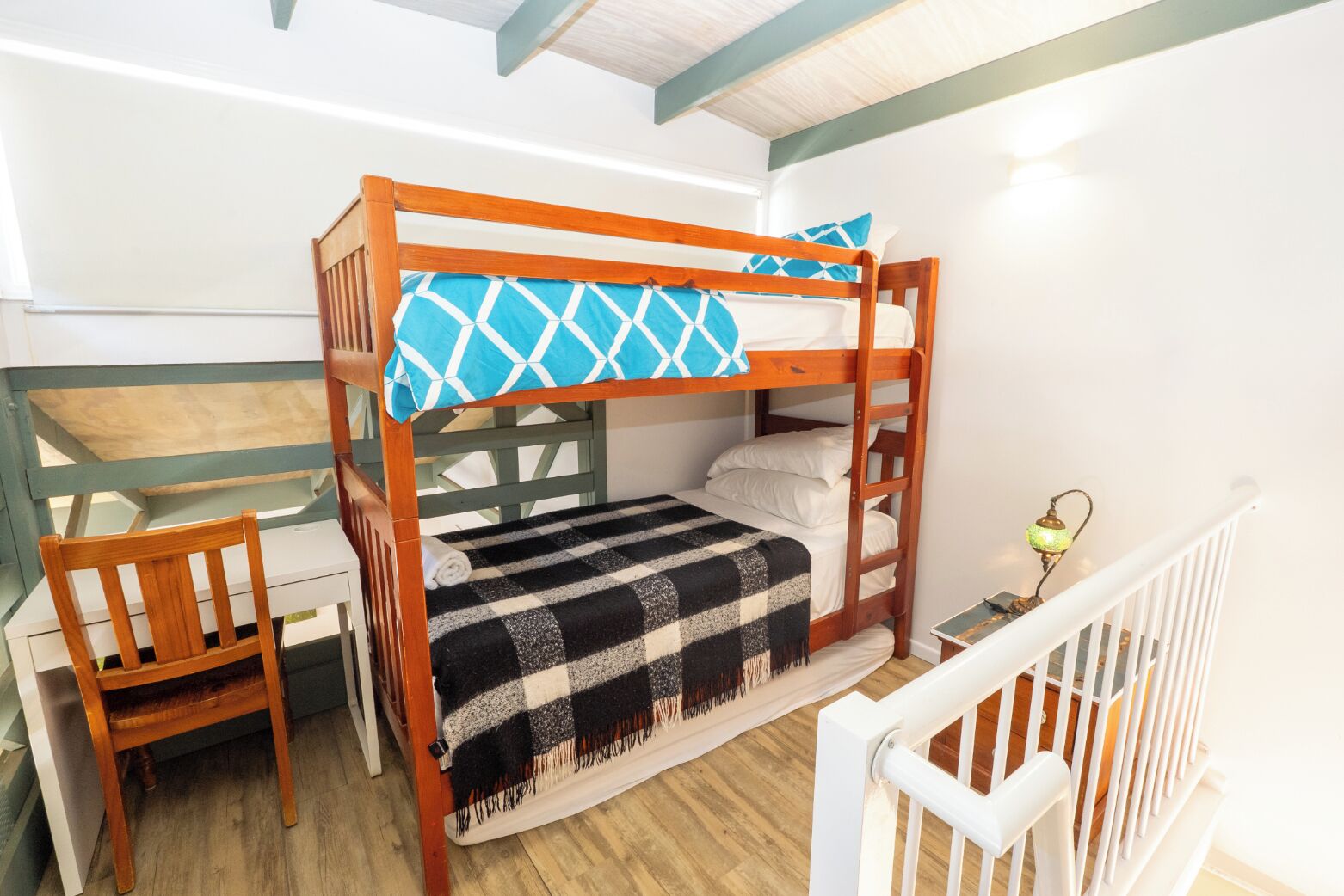 Un lit superposé avec une couverture à carreaux, une chaise en bois et un bureau avec une lampe.