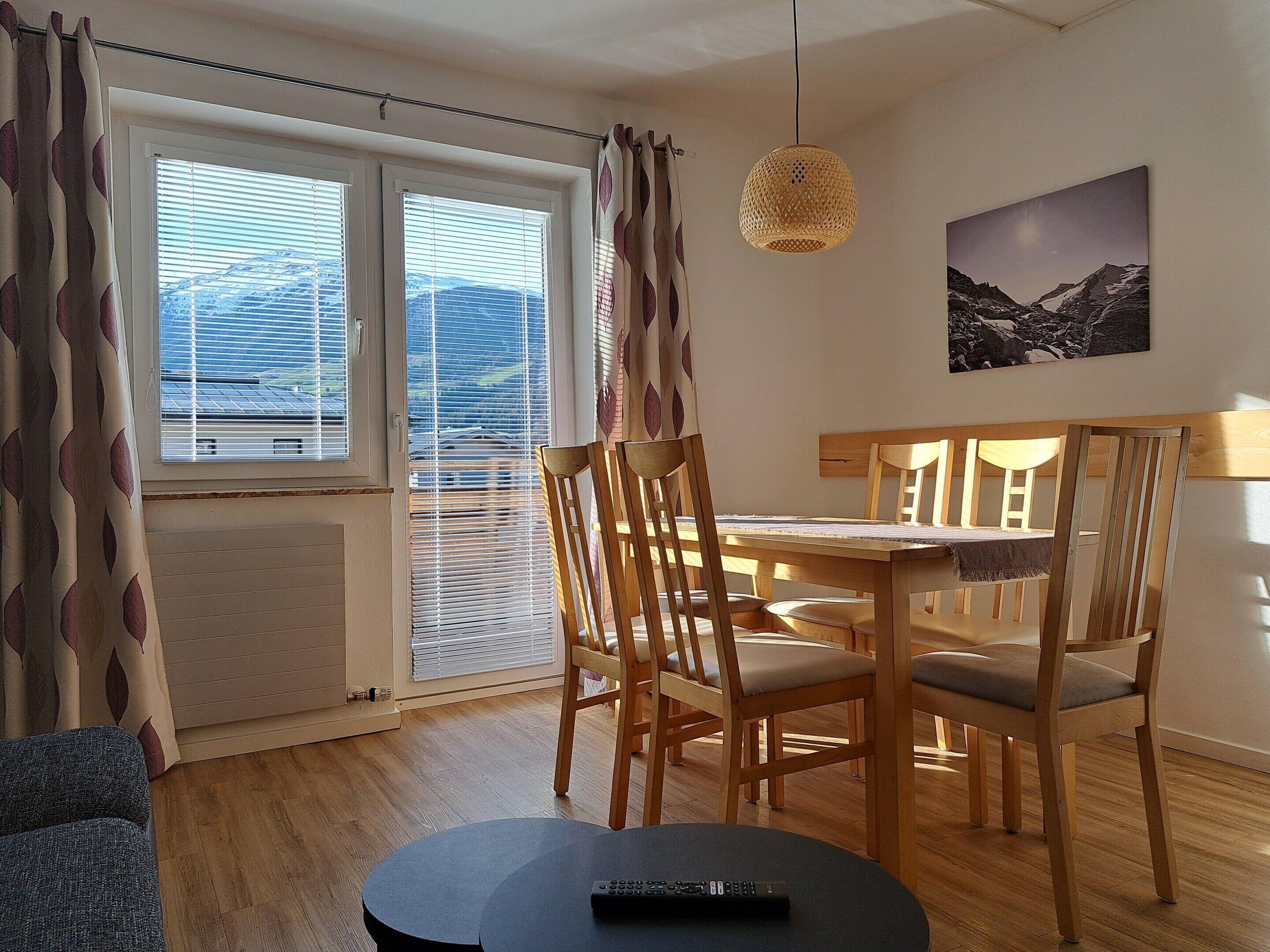 A dining area with a table and chairs, a hanging light fixture, a painting on the wall, and a view of mountains through the window.