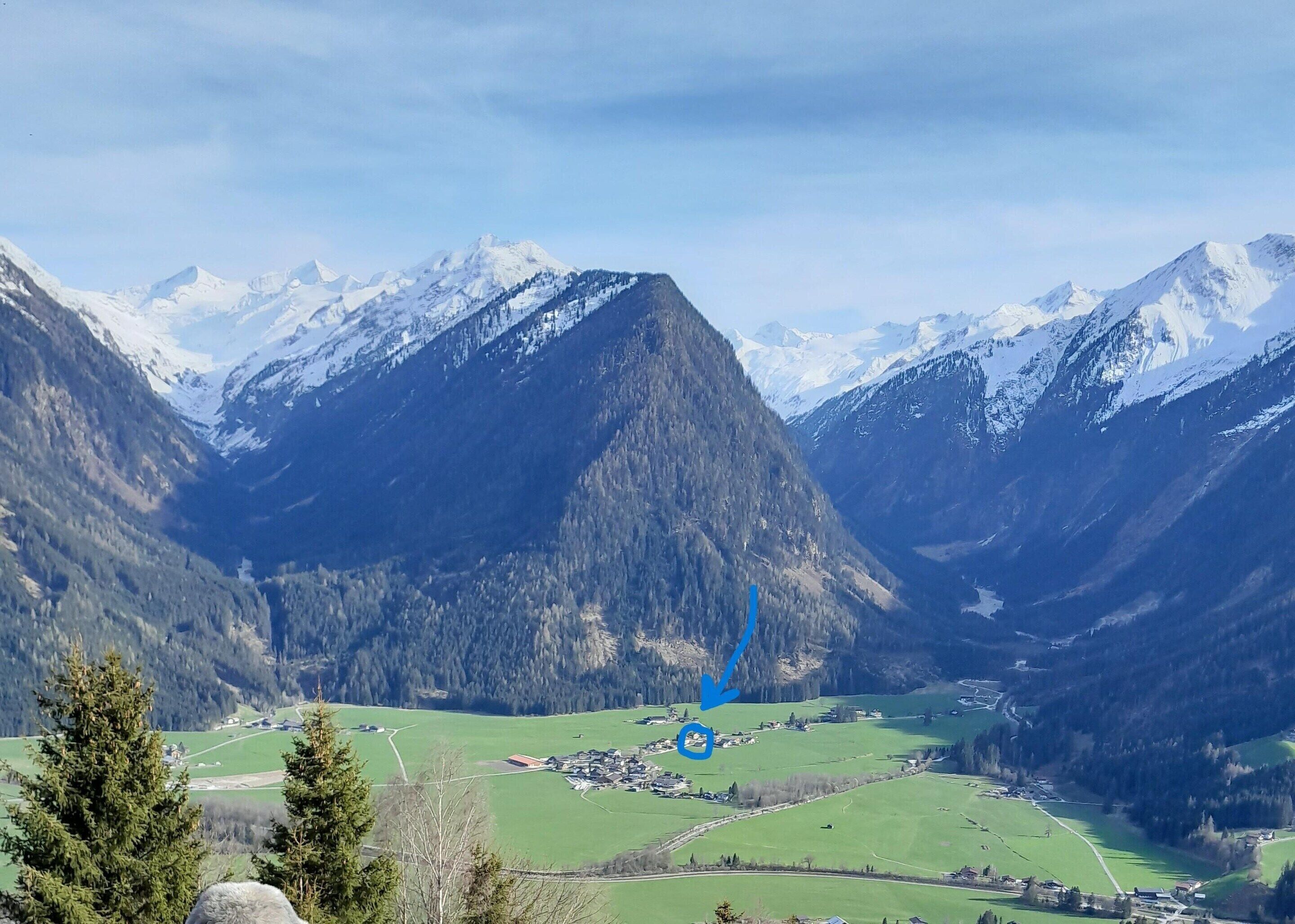 A mountainous landscape with a valley, a small settlement, and snow-capped peaks.