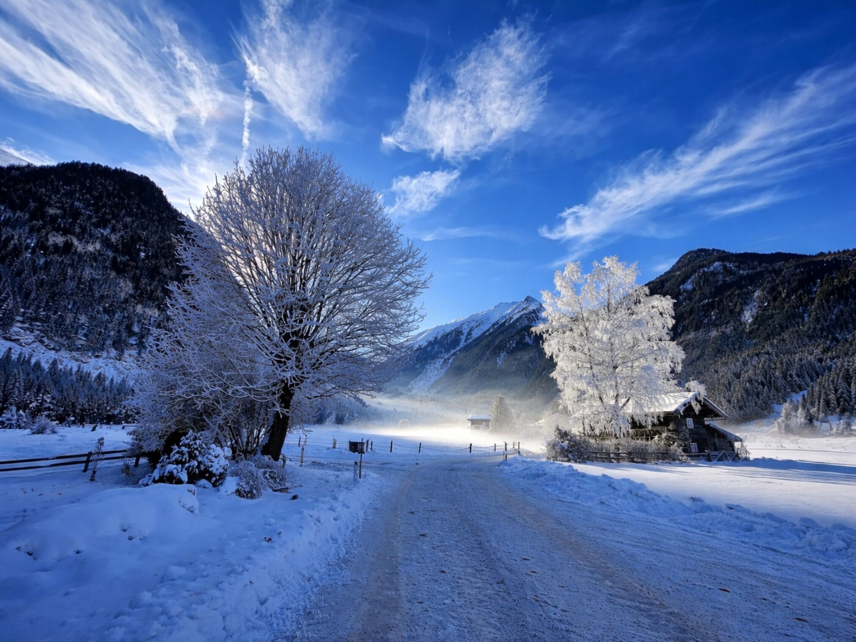 A snow-covered landscape with a clear sky, a wooden cabin, and a mountain in the background.