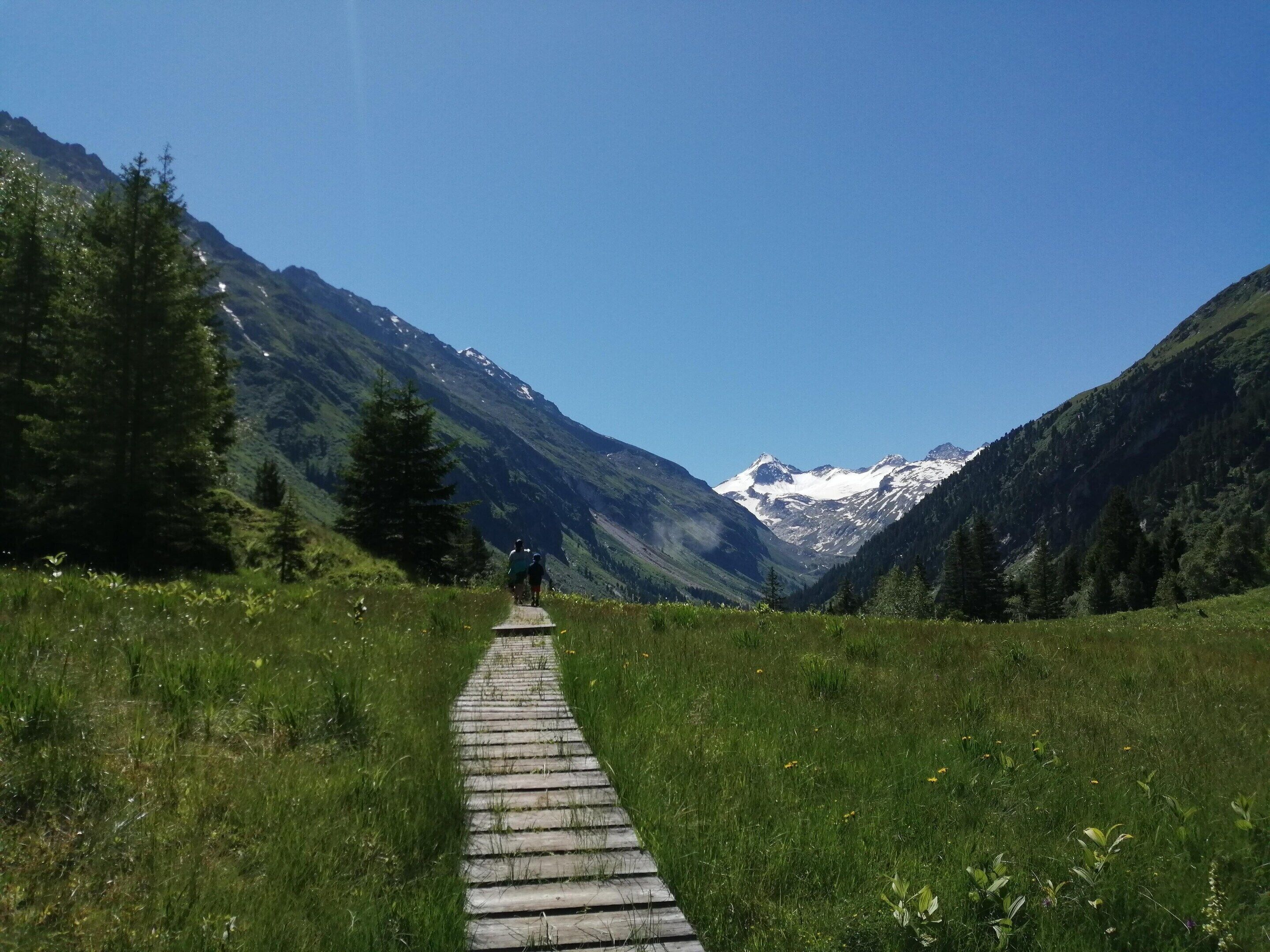 Ein hölzerner Pfad schlängelt sich durch eine gebirgige Landschaft mit schneebedeckten Gipfeln in der Ferne.