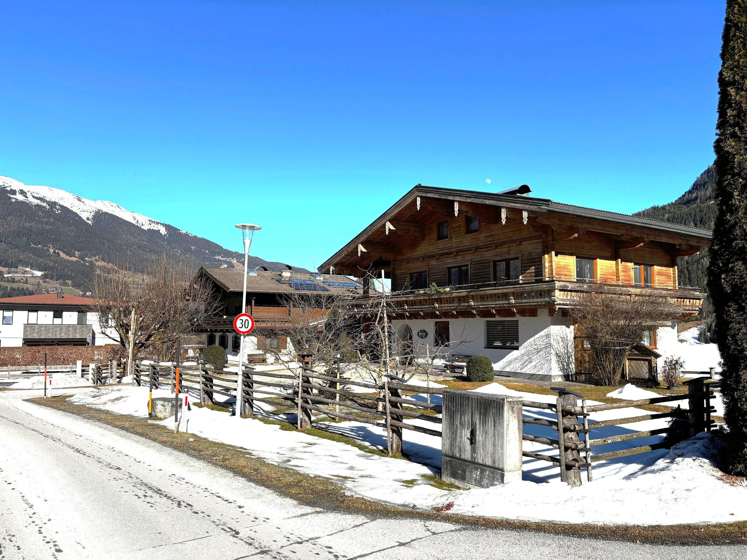 A wooden chalet with a balcony, a mountain backdrop, and a snow-covered ground.