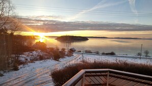 Eine Holzterrasse mit Geländer, die auf eine verschneite Landschaft und einen Sonnenuntergang über dem Wasser blickt.