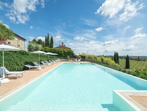 Una piscina con sillones de playa y sombrillas, rodeada de vegetación y vistas al campo.