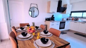Un comedor moderno con una mesa de madera dispuesta para cuatro personas, rodeada de sillas color naranja. Un centro de mesa con frutas se encuentra sobre la mesa, y hay un espejo en la pared.