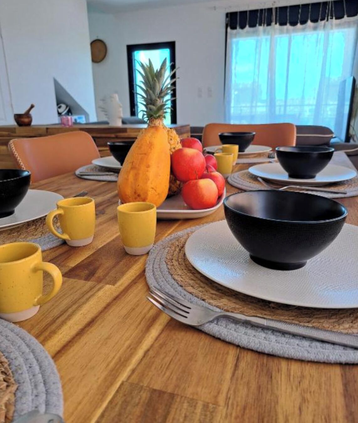 A dining table set with yellow mugs, black bowls, and plates with fruits like pineapple and peaches.