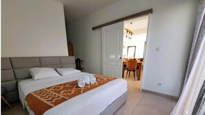 Una habitación de hotel con una cama grande, un área de comedor visible a través de una puerta abierta y una lámpara de araña moderna.