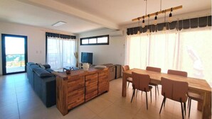 Una sala de estar moderna con un sofá, una mesa de comedor de madera con sillas, un televisor y un ventanal con cortinas.