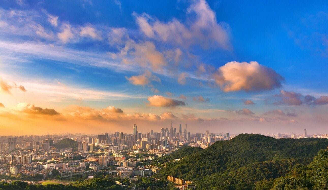 Paisagem urbana com área densa, colina com vegetação e céu claro com nuvens.