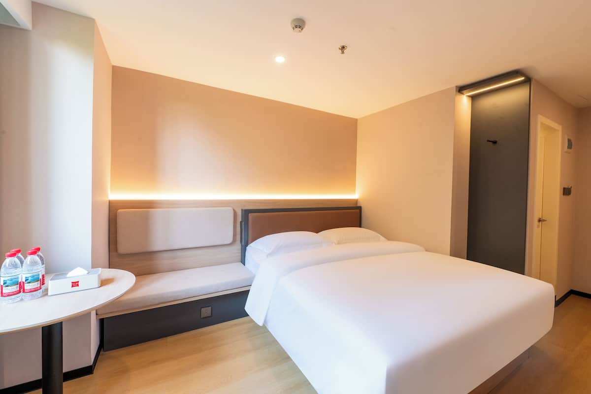 Ein Hotelzimmer mit einem Bett, einem Nachttisch und einer Bank. Auf dem Tisch stehen zwei Wasserflaschen und eine Packung Taschentücher.